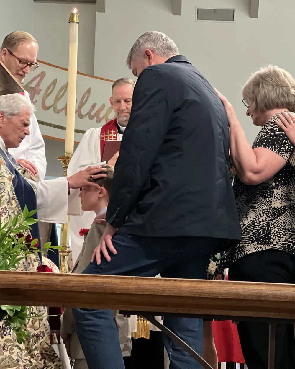 A bishop laying his hand on a young person's head at the altar rail during a confirmation, the family gathered close around them.