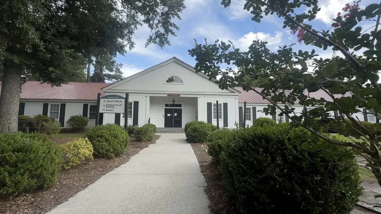 The front of St. Paul's Anglican Church in Summerville: a white clapboard building with a columned portico and a red metal roof, seen along the walkway between clipped hedges under a bright, partly clouded sky.
