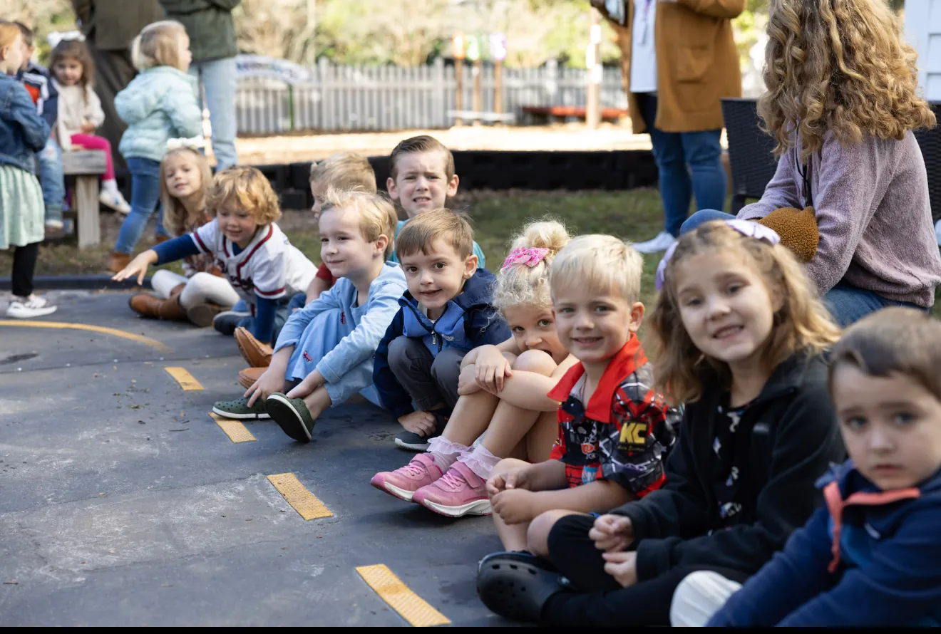 Children sitting in a long row along the edge of the playground at Trinity Classical Academy on a bright morning.