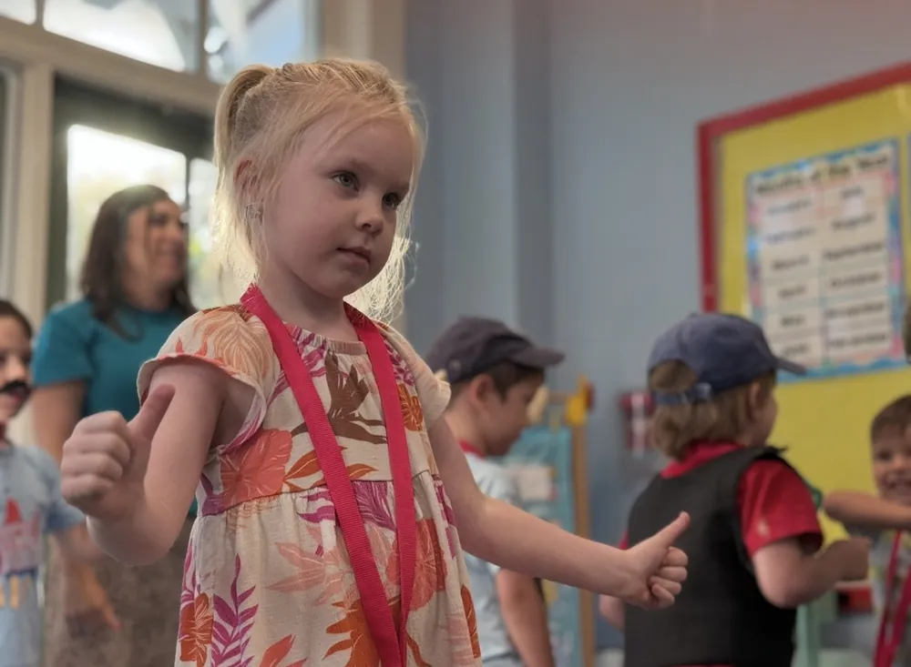 A young girl in a classroom at Trinity Classical Academy giving a thumbs up, classmates and a teacher behind her.